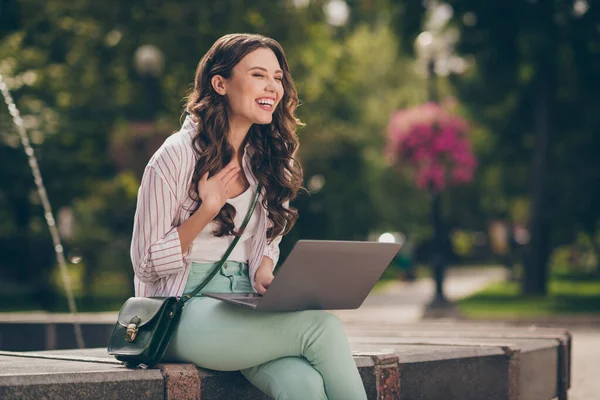 Foto de adorável senhora morena engraçada sentar perto da fonte falando no laptop rindo desgaste pastel jeans camisa bolsa de ombro superior no parque ao ar livre — Fotografia de Stock