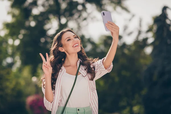 Photo de mignon joyeux cheveux longs ondulés jeune femme faisant selfie show v-sign profiter de la promenade dans le parc porter chemise rayée à l'extérieur — Photo