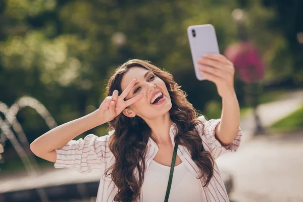 웃고 있는 웃긴 갈색 곱슬머리젊은 여자가 셀피 쇼 v-sign 를 하는 사진 주말 공원에서 줄무늬 셔츠를 입고 즐길 수있다. — 스톡 사진