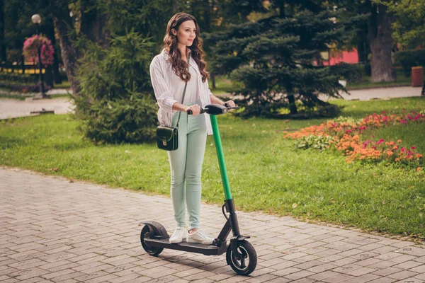 Teljes test fotó csinos fiatal nő göndör barna haj viselet alkalmi ruhát lovagolni meghajtó elektromos robogó eco a parkban — Stock Fotó