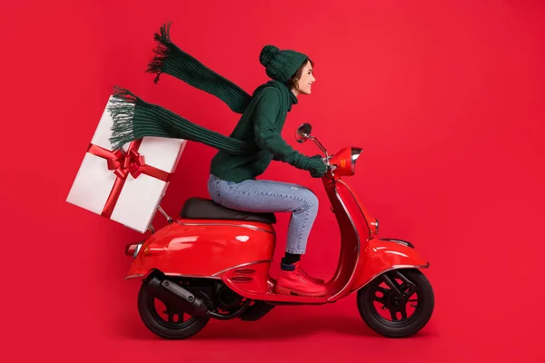 Zijprofiel full size portret van dame rijden bromfiets leveren x-mas pakket jeans laarzen geïsoleerd op rode kleur achtergrond — Stockfoto