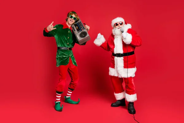 Foto von zwei Personen Elfe halten Blockflöte Show Hörner Santa halten Mikrofon tragen X-mas Kostüm Sonnenbrille Mütze isoliert rote Farbe Hintergrund — Stockfoto