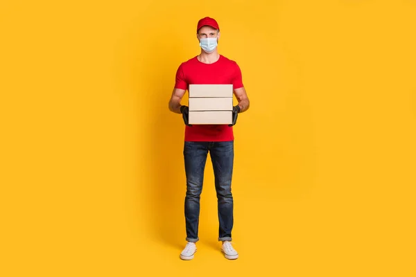 Фото молодого кур'єра, який тримає пальові коробки, носить маску червона футболка капелюх джинсового взуття ізольований жовтий кольоровий фон — стокове фото