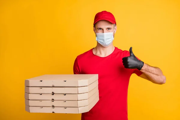 Фото молодого кур'єра, який тримає коробки для піци, піднімають великий палець вгору вдягати маску червона футболка шапка ізольований жовтий колір фону — стокове фото