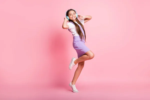 Foto de larga duración de niña pequeña y positiva escuchar música de auriculares inalámbricos aislados sobre fondo de color rosa — Foto de Stock