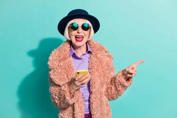 Foto de anciana vestida ropa de abrigo vintage sosteniendo gadget apuntando espacio vacío aislado color turquesa fondo — Foto de Stock