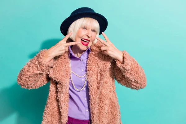 Foto van grappige funky senior dame dragen armen retro jas hoofddeksel tonen v-teken geïsoleerde teal kleur achtergrond — Stockfoto