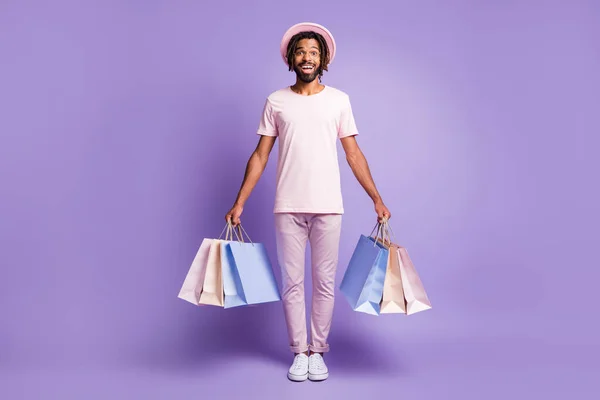 年轻的非洲佬兴奋地穿着粉色T恤衫紫色裤子的男人拿着紫色背景的购物袋的全副照片 — 图库照片