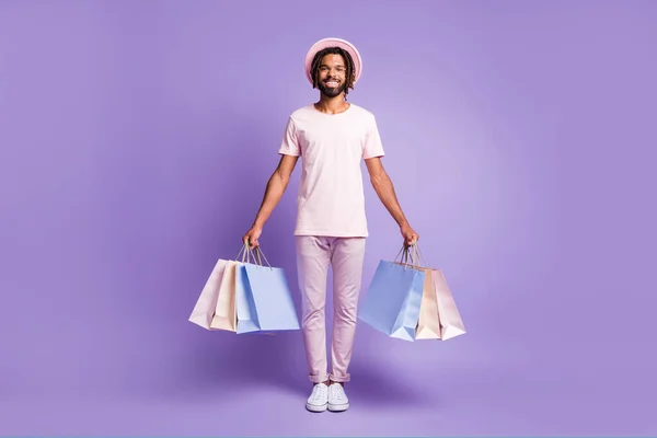 Foto de tamaño completo de joven afro sonriente hombre positivo usar camiseta rosa pantalones violeta mantener bolsas de compras aisladas en el fondo de color púrpura — Foto de Stock