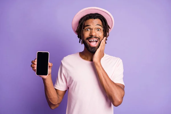 Fotografie mladého afrického vzrušený muž chlap nosit růžové tričko a brýle show telefon displej držet ruku tvář izolované na fialovém pozadí — Stock fotografie