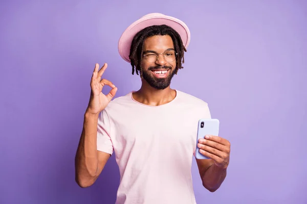 Foto portrét mrkajícího muže ve stylovém klobouku čepice čelenky zobrazující ok-sign drží telefon v jedné ruce izolované na živém fialové barevné pozadí — Stock fotografie