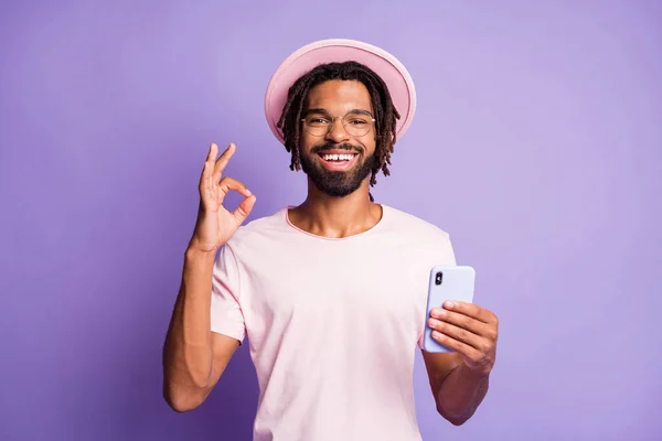 Portrait photo de l'homme en chapeau chapeau élégant coiffure montrant ok-signe tenant le téléphone dans une main isolée sur fond violet vif — Photo