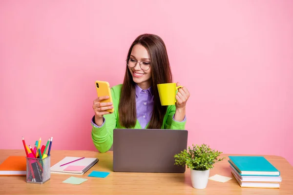 Menina positiva estudante do ensino médio sentar mesa uso smartphone segurar caneca desgaste camisa verde isolado cor rosa fundo — Fotografia de Stock