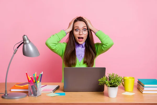 Foto de chica trabajadora asombrada grito toque manos cabeza desgaste camisa verde sentarse mesa aislado pastel color fondo — Foto de Stock