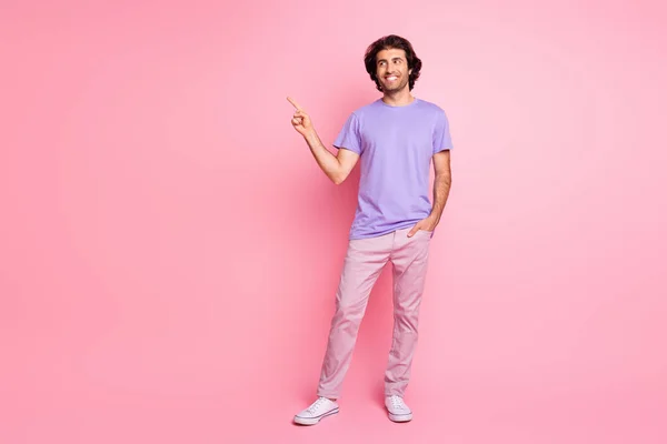 Full length photo of man indicate finger look empty space wear purple shirt pants sneakers isolated pastel pink color background