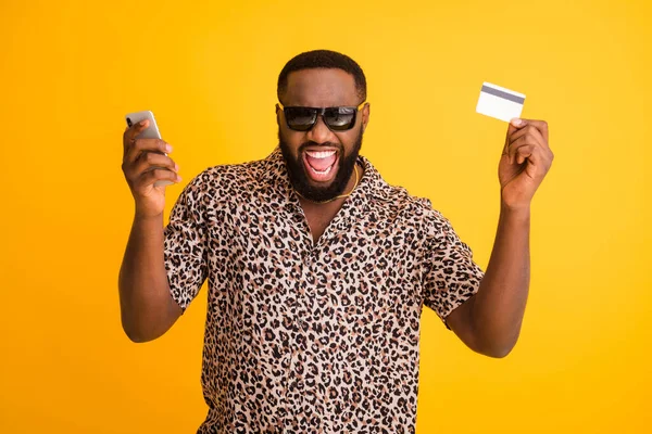 Foto de loco divertido chico de piel oscura mantener el teléfono hacer el pago en línea transferencia de compra uso de tarjeta de crédito usar gafas de sol collar de oro camisa de leopardo aislado de color amarillo brillante fondo — Foto de Stock