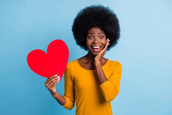 Retrato fotográfico de la feliz niña de piel negra sonriente mostrando la tarjeta de corazón de papel rojo tocando la mejilla aislada en un fondo de color azul brillante —  Fotos de Stock