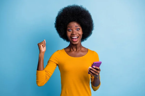 Foto portrét ohromený černošky kůže dívka gestikulace jako vítěz vedení smartphone izolované na jasně modré barvy pozadí — Stock fotografie