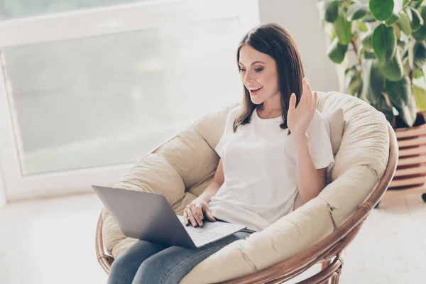 Foto gadis video chatting di laptop melambaikan tangan mengenakan pakaian gaya kasual duduk kursi wicker di rumah di dalam ruangan — Stok Foto