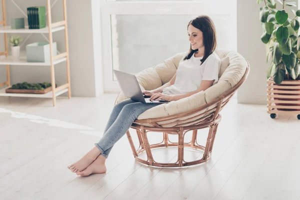 Фотография в полный рост, на которой барышня сидит на плетеном стуле, работающем на ноутбуке, в белой футболке с джинсами в доме внутри — стоковое фото