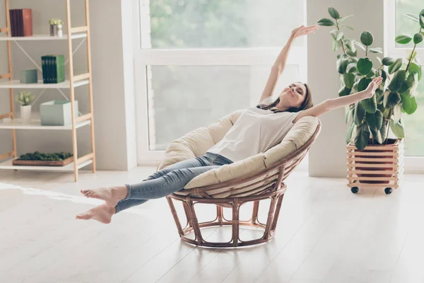 Foto de tamaño completo de niña positiva sentarse silla de mimbre brazos estirados desgaste ropa de estilo casual descalzo en casa en interiores — Foto de Stock