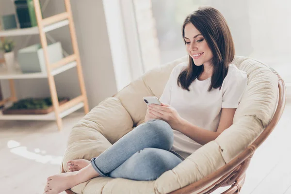 긍정적 인 소녀가 앉는 의자 사진 스마트폰에서 소셜 미디어 뉴스를 읽고 방에서 평상복을 입고 — 스톡 사진