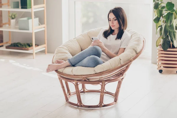 Full size foto van charmant meisje zitten rieten stoel chatten met vrienden op smartphone in huis — Stockfoto