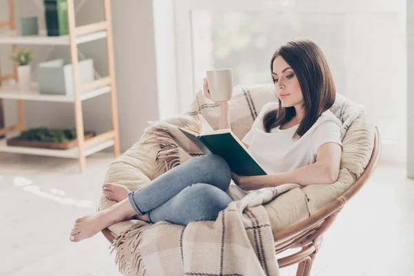 Foto de longitud completa de la niña concentrada sentarse en la silla de mimbre leer libro celebrar taza de café en la habitación de la casa moderna — Foto de Stock