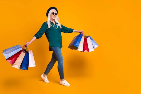 Profilo completo del corpo foto di funky nonna signora camminare centro commerciale portare molti pacchetti indossare camicia verde sole specifiche collana jeans scarpe cappuccio retrò isolato luminoso colore giallo sfondo — Foto Stock