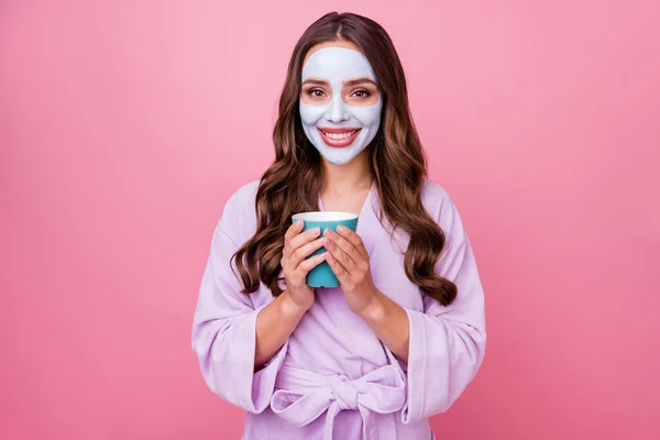 ピンク色の背景に隔離されたバスローブを着てコーヒーのカップを保持顔マスクを身に着けている皮膚のための治療手順を行うかなりかわいい女の子の写真 — ストック写真
