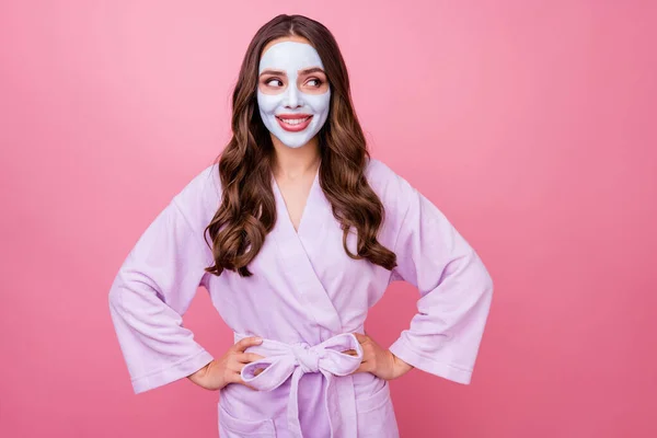 Foto retrato de chica bonita con máscara facial largo pelo castaño ondulado mirando a un lado manteniendo las manos en la cintura sonriendo aislado en el fondo de color rosa —  Fotos de Stock