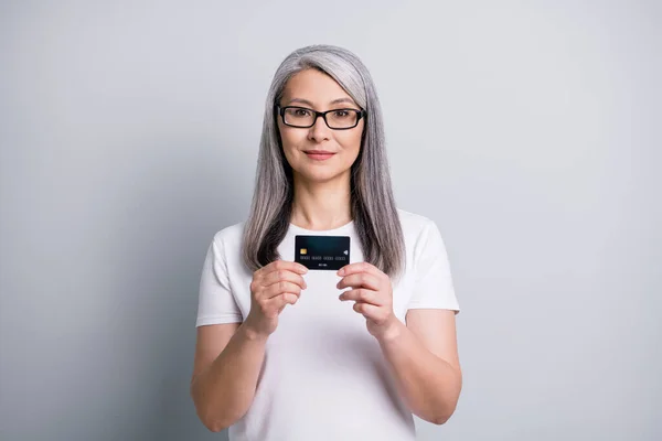 Foto de pensionista anciana celebrar la presentación de plástico tarjeta de crédito desgaste especificaciones blanco camiseta aislado color gris fondo —  Fotos de Stock
