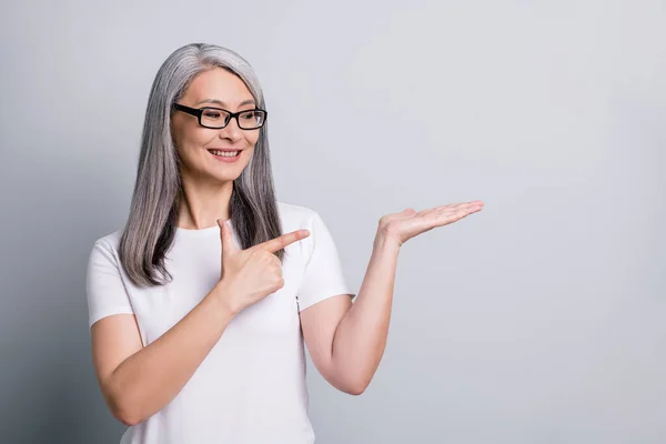 Foto de anciana indican brazo de sujeción del dedo mirada espacio vacío usar gafas camiseta blanca aislado fondo de color gris — Foto de Stock