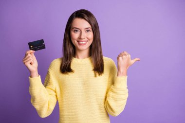 Photo portrait of pretty girl holding bank plastic card showing with thumb on blank space smiling isolated on bright purple color background clipart