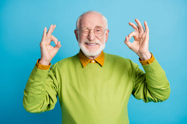 Foto de anciano retirado levantar dos manos mostrar okey guiño ojo desgaste gafas verde jersey aislado color azul fondo — Foto de Stock