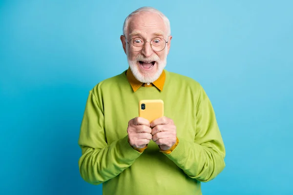 Фото пенсионера дедушки держать телефон смотреть экран рот открытый носить спецификации зеленый пуловер изолированный синий цвет фона — стоковое фото