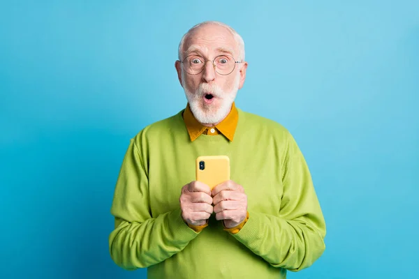 Photo de retraité vieux tenir téléphone cellulaire expression choquée porter des lunettes pull vert isolé fond de couleur bleue — Photo