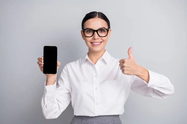 Foto av ganska promotor flicka hålla telefonen skärmen tomt utrymme höja tummen upp slitage specs vit skjorta isolerad grå färg bakgrund — Stockfoto