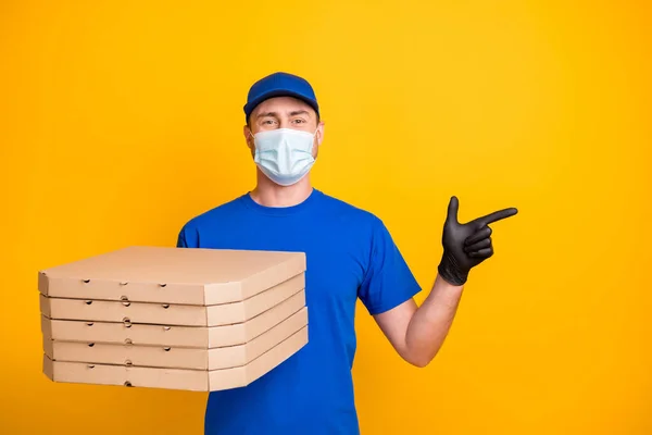 Retrato de entregador indicam dedo espaço vazio segurar pizzas usar cuidados de saúde máscara facial isolado no fundo cor amarela — Fotografia de Stock