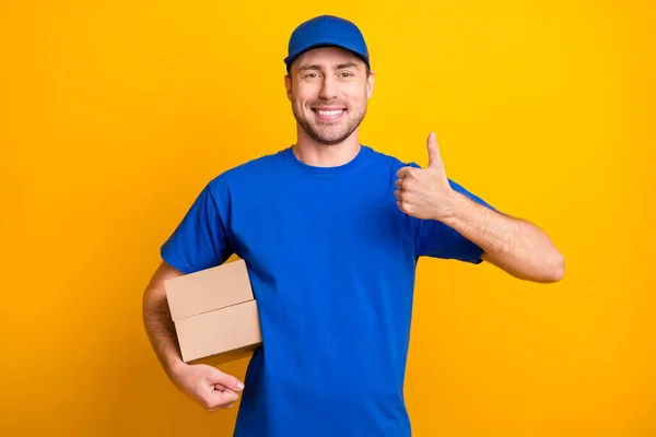 Foto von positiven Kerl zeigen Daumen nach oben Arm halten Karton Paket tragen Hut isoliert auf gelbem Hintergrund — Stockfoto