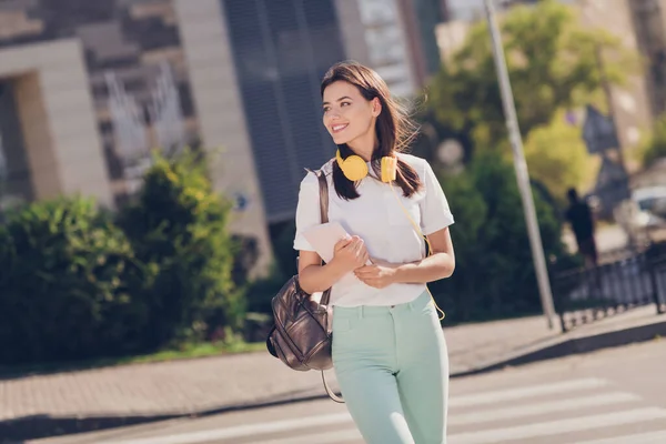 Foto retrato de mulher cruzando rua segurando Copybook com fones de ouvido amarelos ao redor do pescoço ao ar livre — Fotografia de Stock