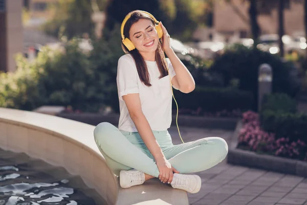 Foto retrato de una mujer sentada en pose de loto en el borde de la fuente escuchando música tocando auriculares amarillos con una mano en el parque — Foto de Stock
