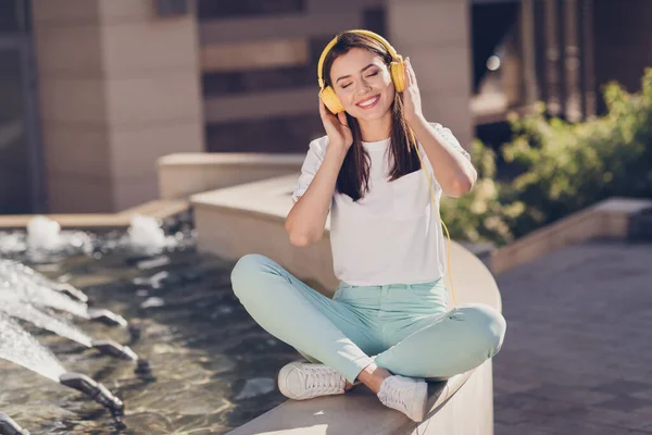 Foto retrato de niña sentada en pose de loto en el borde de la fuente escuchando música tocando auriculares amarillos con las manos al aire libre — Foto de Stock