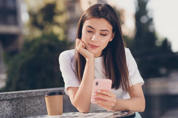 Foto retrato de una niña sentada en la mesa con una taza de papel sosteniendo la cabeza de la barbilla del teléfono con una mano al aire libre — Foto de Stock