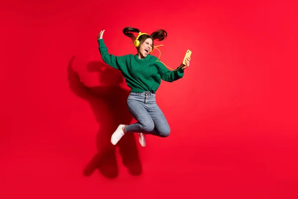 Full length φωτογραφία του αξιολάτρευτο νεαρή κοπέλα φορούν πράσινο πλεκτό πουλόβερ ακούγοντας μουσική ακουστικά χορό άλμα απομονωμένο κόκκινο χρώμα φόντο — Φωτογραφία Αρχείου