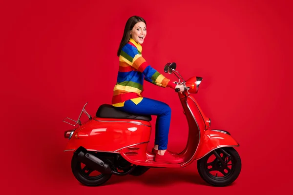 Профільний вид збоку досить весела дівчина, що їде на мопеді швидка швидкість, розважається ізольовано на яскраво-червоному кольоровому фоні — стокове фото