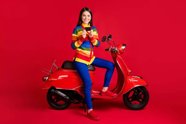 Foto retrato de menina segurando cartão de crédito em duas mãos sentado em scooter isolado no fundo colorido vermelho vívido — Fotografia de Stock