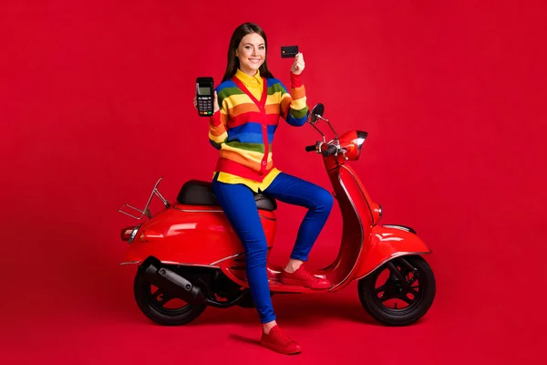 Foto retrato de mulher feliz segurando terminal de cartão de crédito em mãos sentadas em scooter isolado em fundo colorido vermelho vívido — Fotografia de Stock