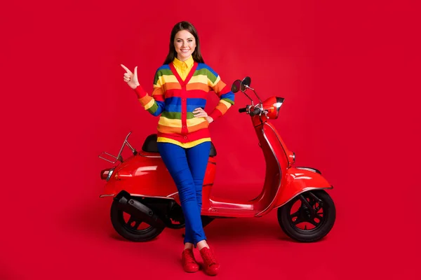 Foto retrato de mulher apontando dedo para o lado sentado em scooter isolado no fundo colorido vermelho vívido — Fotografia de Stock