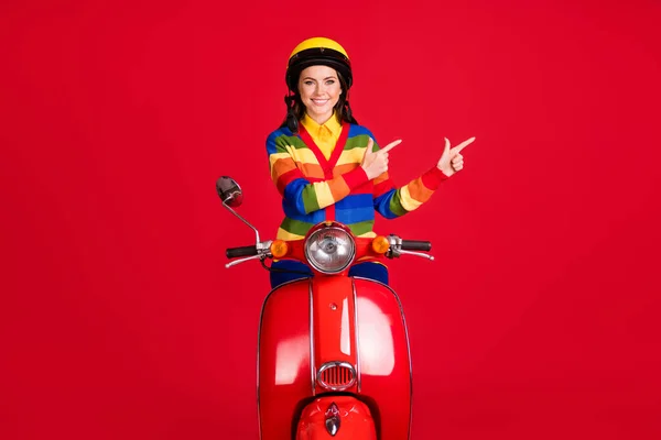 Foto portret van lachend meisje wijzen twee vingers naar kant rijden scooter geïsoleerd op levendige rood gekleurde achtergrond — Stockfoto